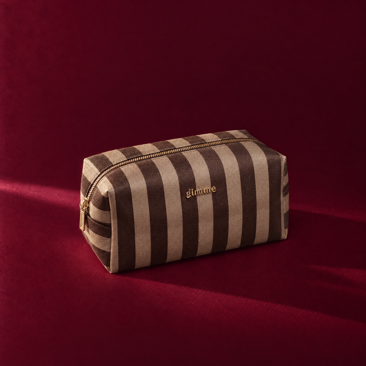 Gestreepte bruin-beige pouch met gouden rits op een bordeauxrode achtergrond. Gratis pouch bij bestellingen vanaf €50 bij Gioia store.