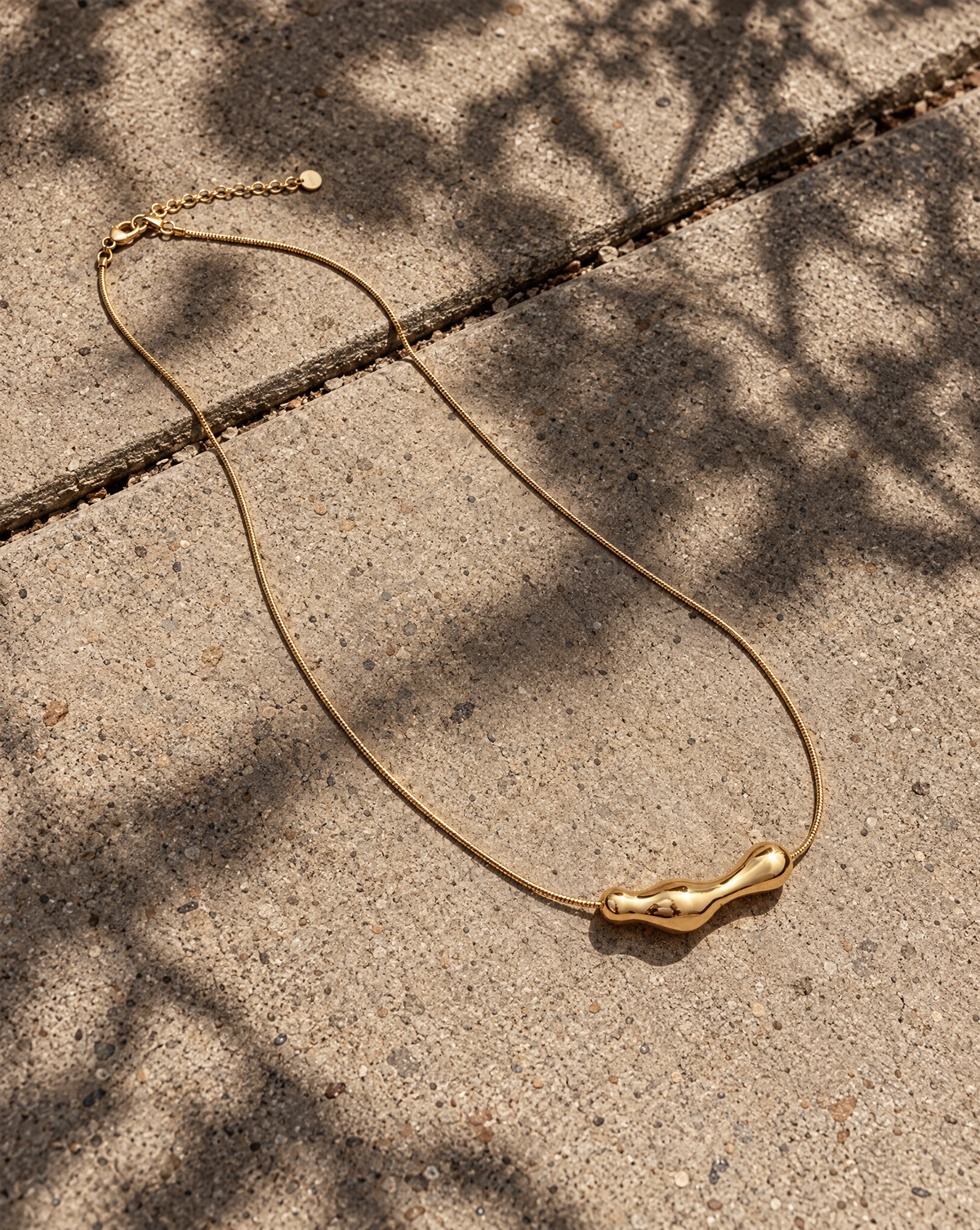 Minimalistische Aura Necklace goudkleurige RVS ketting met organische hanger op beton.