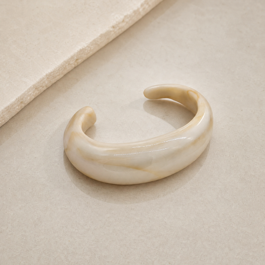 The soft stond bangle in offwhite met een beige marmering erin. Het design is een open bangle dit dikker is in het midden en taps toeloopt richting de einden. Gemaakt van hars, dus lichtgewicht en comfortabel