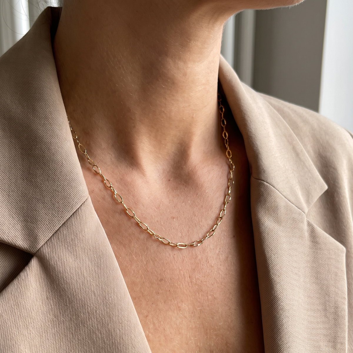 Vrouw draagt de Luna Chain goudkleurige RVS schakelketting met langwerpige schakels en een beige blazer.